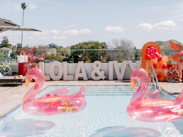 A pool party setup with two pink flamingo floats, large block letters spelling &ldquo;NOLA & IVY&rdquo; by the pool, and pastel decorations in a sunny backyard.