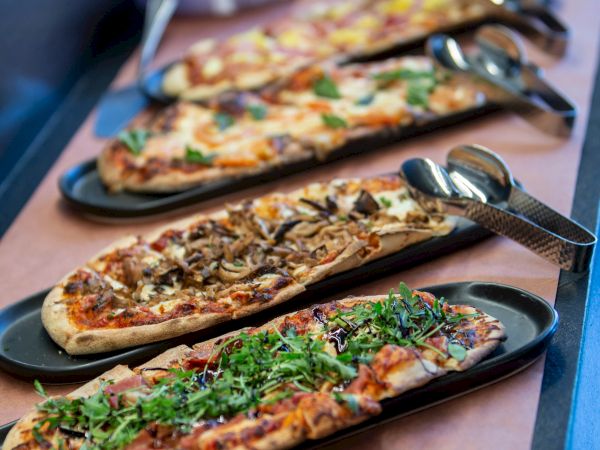 An array of loaded flatbread pizzas topped with cheese, herbs, and vegetables on a serving line, ready to serve.