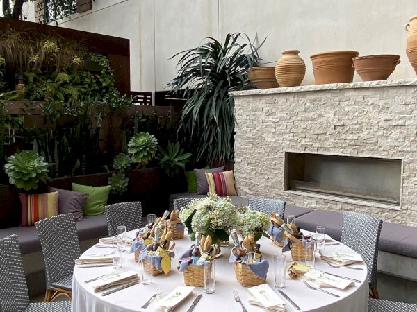Outdoor dining setup with a round table, elegant place settings, and wicker chairs; potted plants and a stone fireplace create a chic terrace vibe.