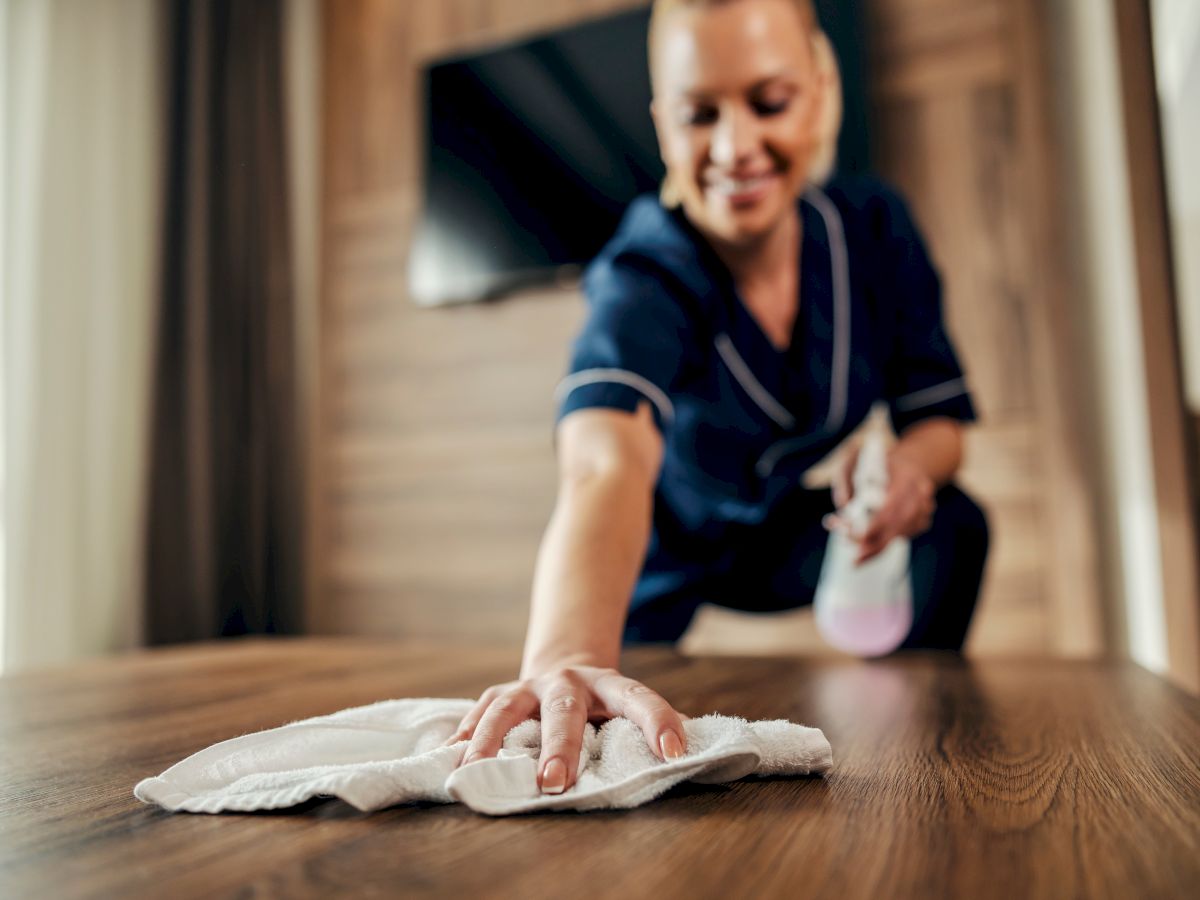 A janitor is cleaning a wooden table, wiping with a cloth while smiling in the foreground; he&rsquo;s wearing a dark uniform in a cozy room.