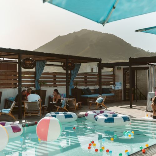 An outdoor pool with colorful inflatable tubes, sun umbrellas, lounge chairs, and people relaxing by a mountain-view deck.