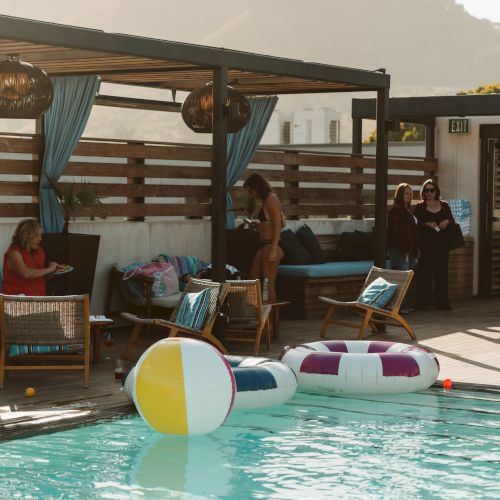Outdoor pool scene with people relaxing under a cabana, lounge chairs, and striped inflatable tubes with mountains in the background.