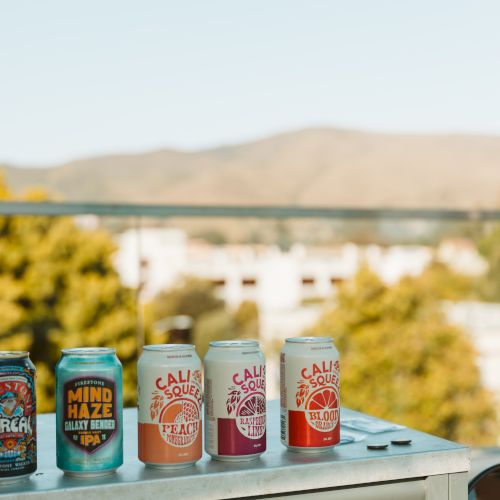 Colorful beverage cans sit on a table outdoors with a blurred landscape and hills in the background.