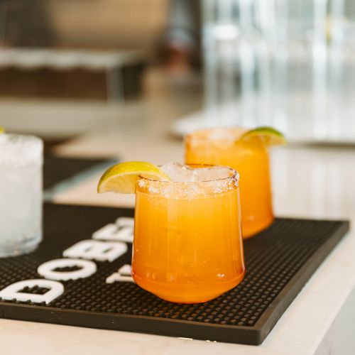 A glass pitcher of orange juice on a tray, with lemon slices and ice, beside a blurred drink container.