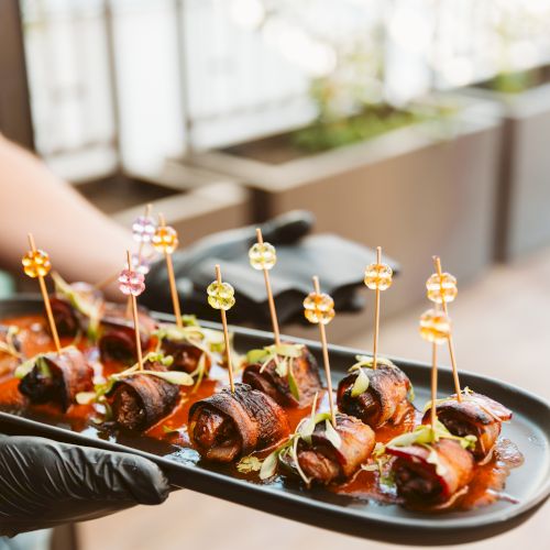 A server holds a platter of colorful skewer-topped grilled bites in sauce, garnished with herbs.