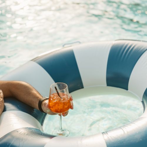 A hand on a white-and-blue inflatable boat holds a glass of iced amber drink while floating on the bright water.