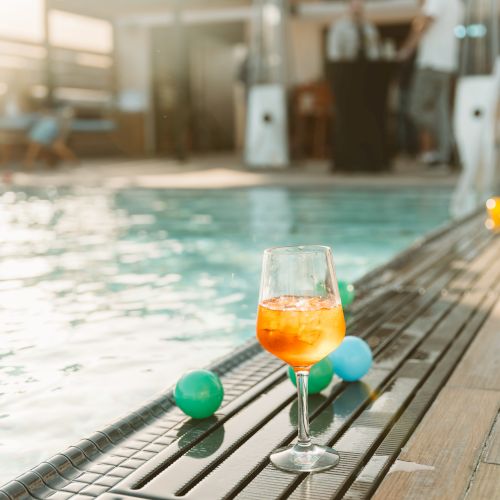 A poolside lounge shows a glass of orange drink with ice, plus green float balls in sunny blur.