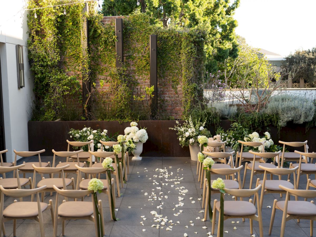 An elegant outdoor event setup with rows of wooden chairs, floral centerpieces, and a lush ivy-covered garden wall.