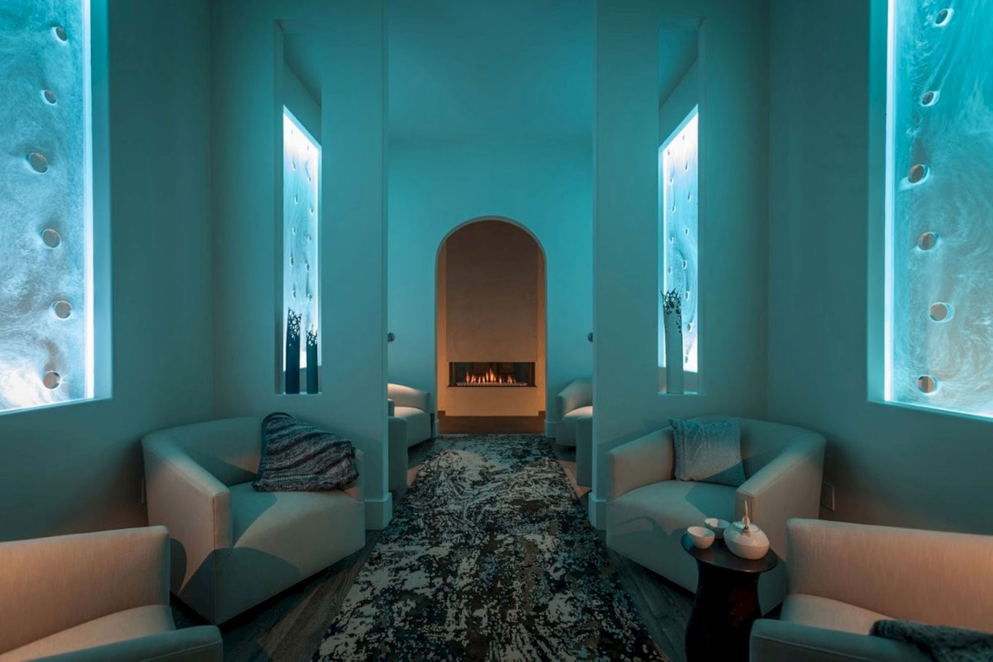 A cozy modern lounge with teal walls, two sofas, a central fireplace glow, framed windows, and a patterned rug.