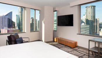 A modern hotel room with a large bed, TV, chair, and stunning city skyline views through multiple windows.