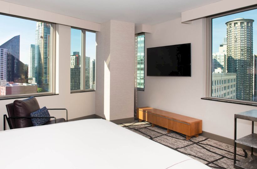 A modern hotel room with a large bed, TV, chair, and stunning city skyline views through multiple windows.