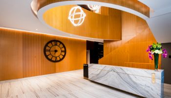 A modern lobby with a marble reception desk, wooden spiral ceiling, large wall clock, and decorative orb lights, featuring floral arrangements.