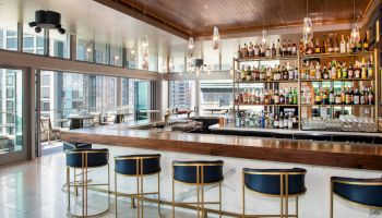 A modern bar features a well-stocked shelf, elegant stools, and large windows with city views, creating a stylish atmosphere.