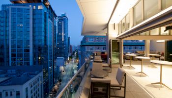 A modern rooftop terrace with seating overlooks city buildings at dusk, featuring a stylish interior space with tables.