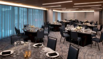 The image shows an elegantly set banquet room with round tables, black chairs, floral centerpieces, and dim lighting.