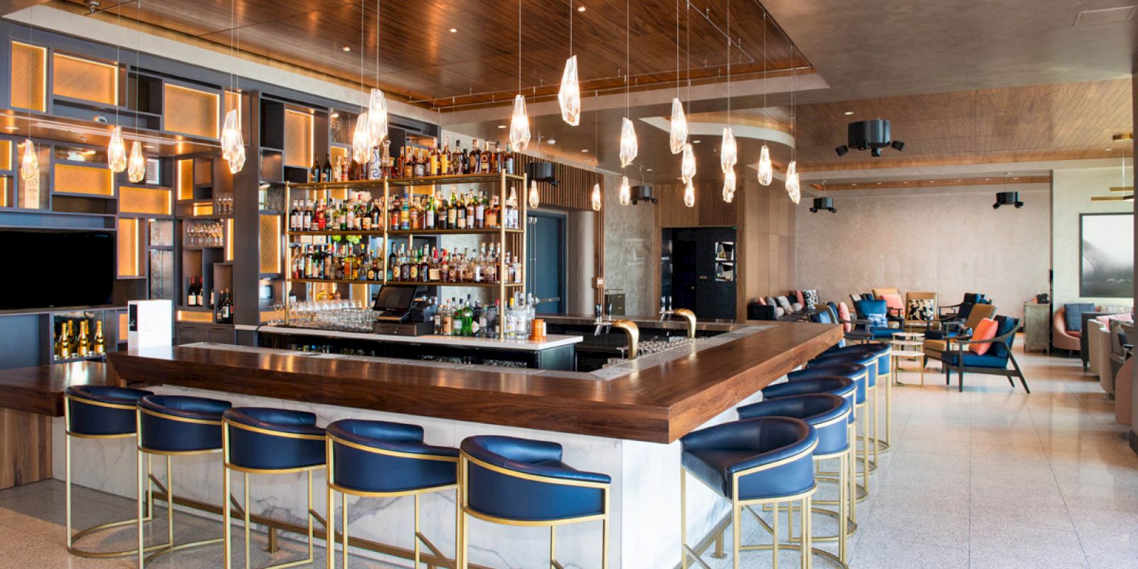 The image shows a modern bar interior with a central counter, blue bar stools, hanging lights, shelves of bottles, and a seating area.