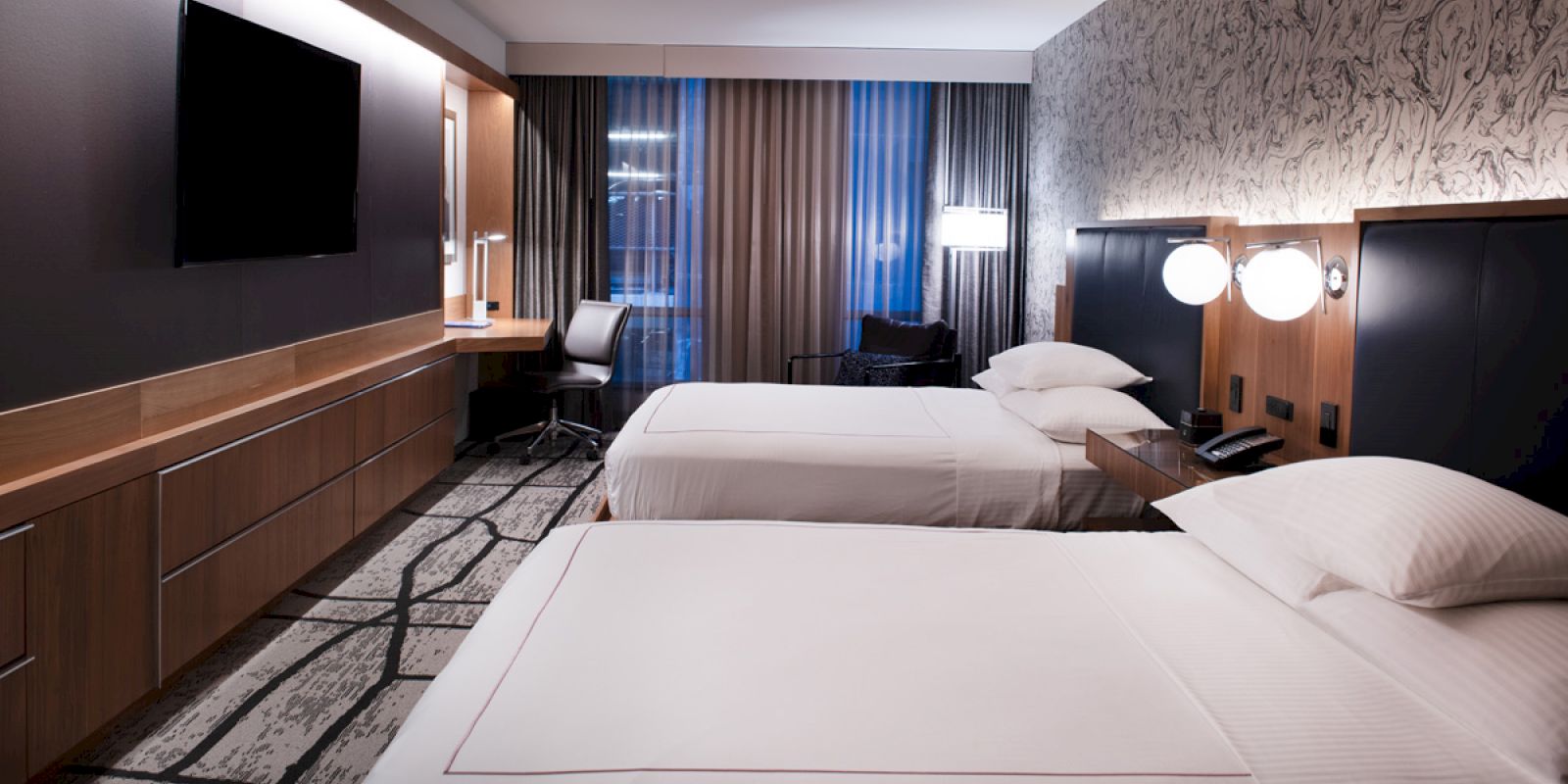 The image shows a modern hotel room with two beds, a large TV, a patterned carpet, and a work desk with a chair by the window.
