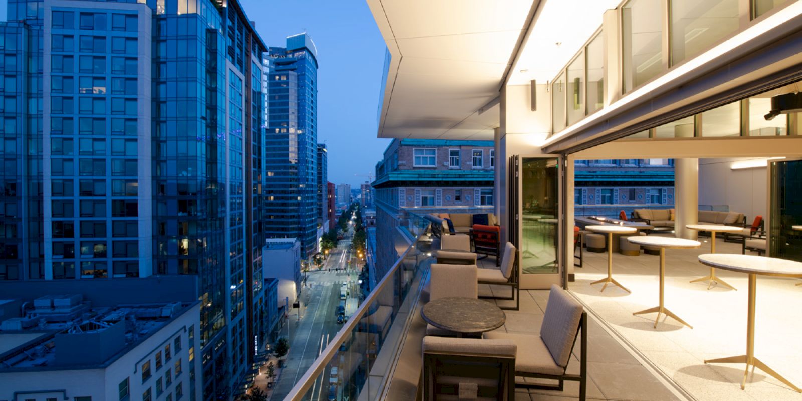 A cityscape view from a modern balcony at dusk, featuring glass buildings, a tiled patio with seating, and interior tables with lighting.