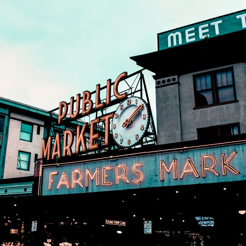 The image shows the iconic neon signs of Pike Place Market, featuring "Public Market" and "Farmers Market" against a cityscape backdrop.