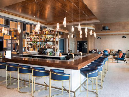 A modern bar with blue stools, hanging lights, and a fully stocked back shelf. There’s a lounge area in the background for seating.