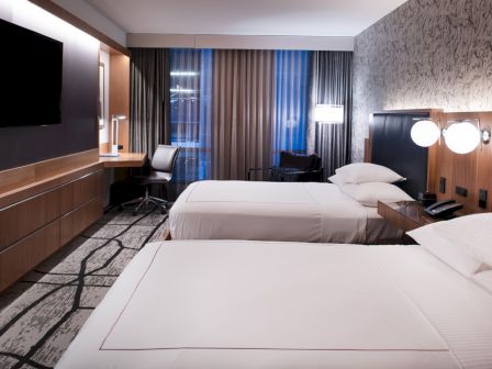 The image shows a modern hotel room with two beds, a flat-screen TV, a desk and chair, wall lighting fixtures, and patterned carpet.