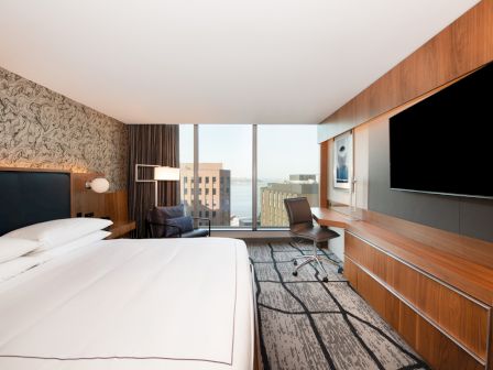 A modern hotel room features a large bed, patterned wallpaper, desk with chair, and a big window offering a city view.