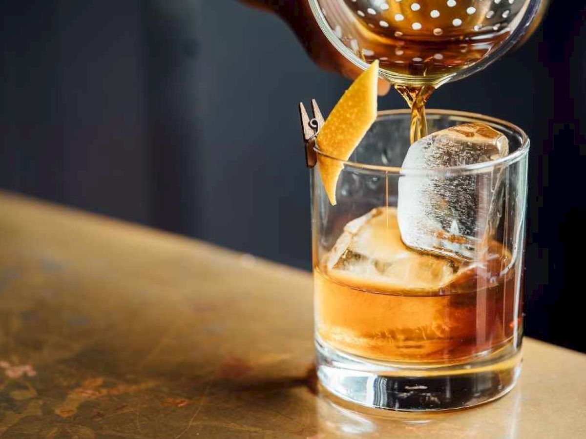 A beverage is being poured into a glass with a large ice cube and an orange twist garnish on a bar counter.