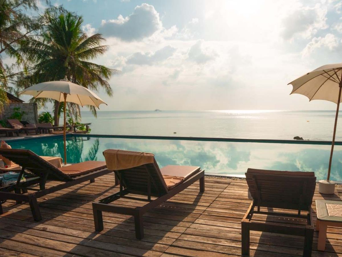 A serene tropical setting with lounge chairs, umbrellas, a pool, and a view of the ocean, framed by palm trees under a sunny sky.