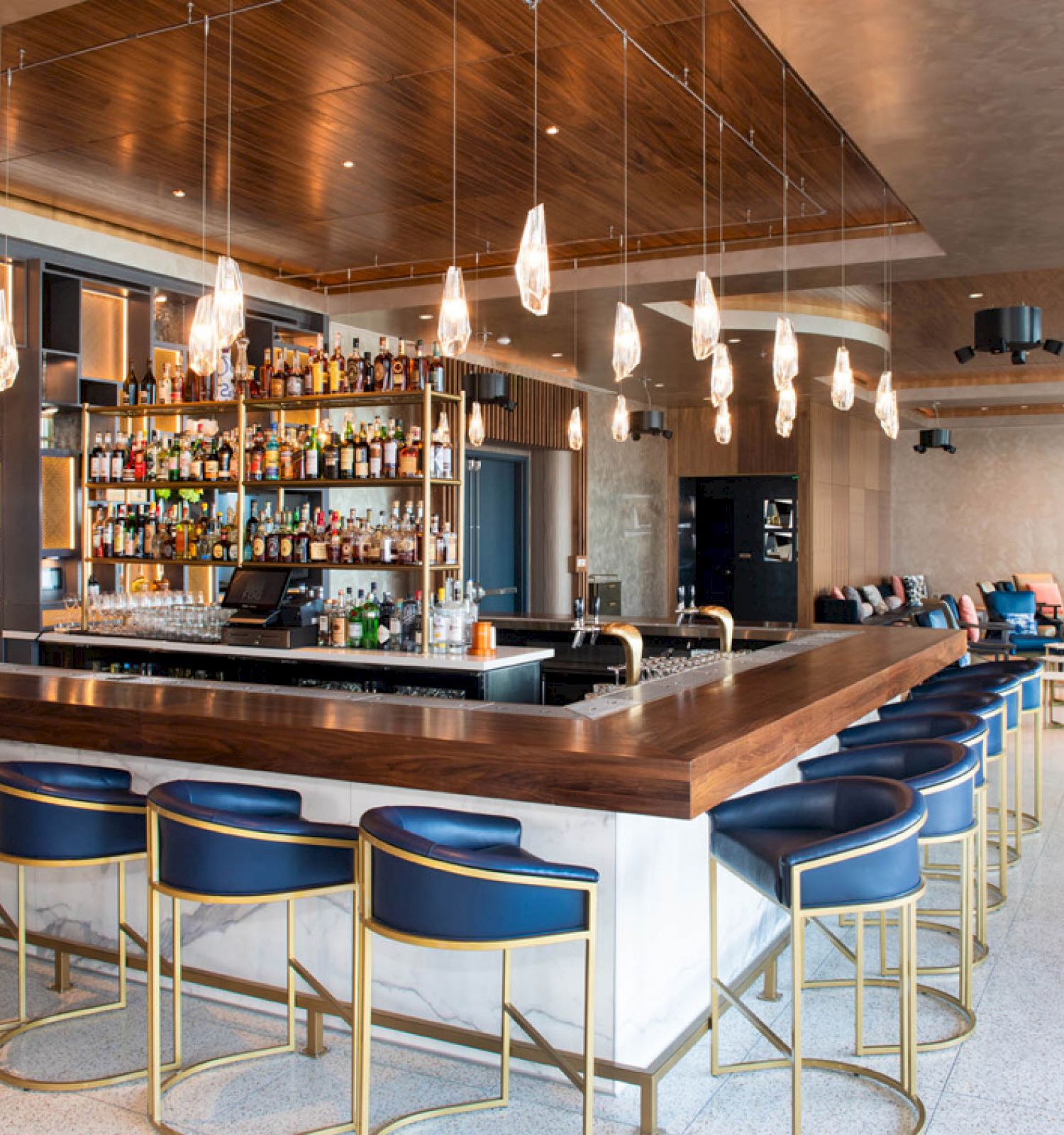 A modern bar with a stocked counter, blue bar stools, pendant lights, and lounge seating in the background, creating a chic ambiance.