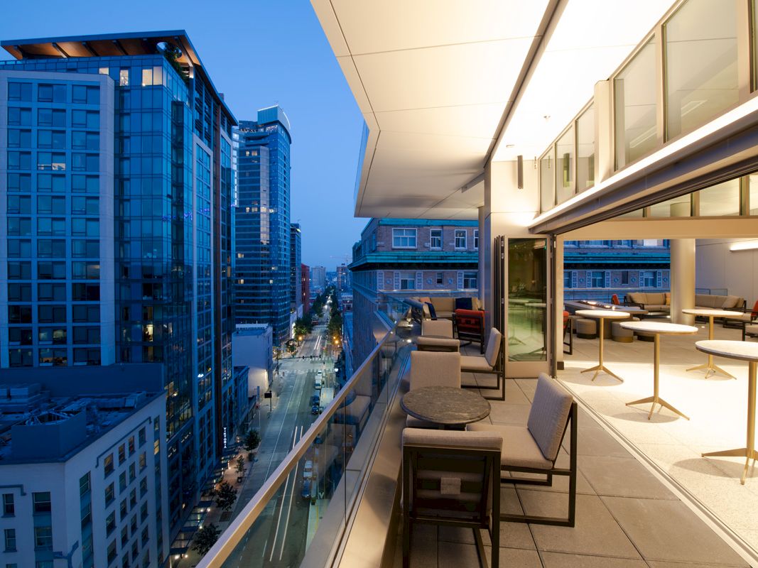A modern cityscape at dusk, featuring a rooftop terrace with tables, chairs, and city views. Skyscrapers line the street below.