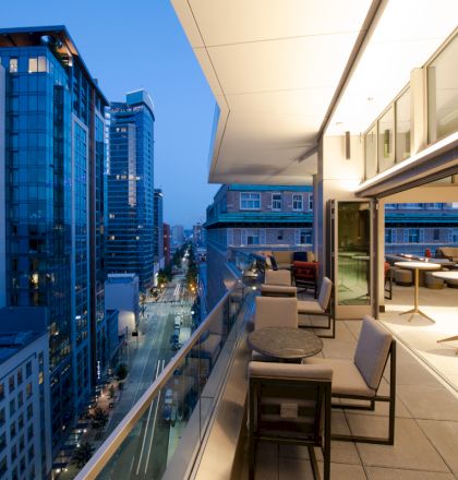 A modern cityscape with high-rise buildings and a stylish balcony overlooking a street, featuring outdoor seating and tables.