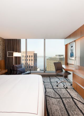 A modern hotel room features a large bed, wall-mounted TV, desk, and window with a city view in the background.