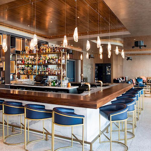A modern bar with stylish stools, hanging lights, and a stocked shelf. Comfortable seating is in the background for guests.