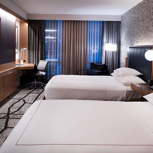 The image shows a modern hotel room with two beds, a wall-mounted TV, a desk, and stylish decor.