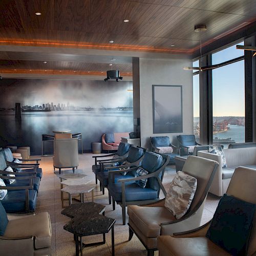 A modern lounge with plush seating, wall art, large windows offering a water view, and natural light creating a cozy atmosphere.
