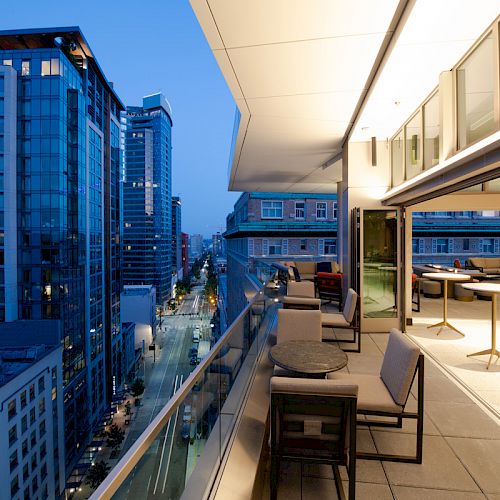 The image shows a modern rooftop terrace with outdoor seating overlooking a cityscape with tall buildings at twilight.