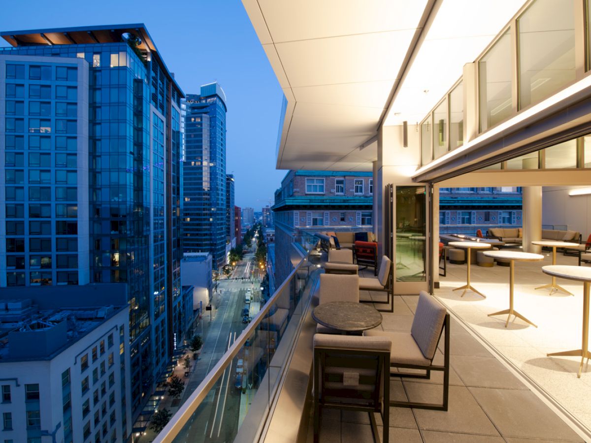 A modern balcony with tables and chairs overlooks a cityscape with tall buildings at dusk under a clear, blue evening sky.