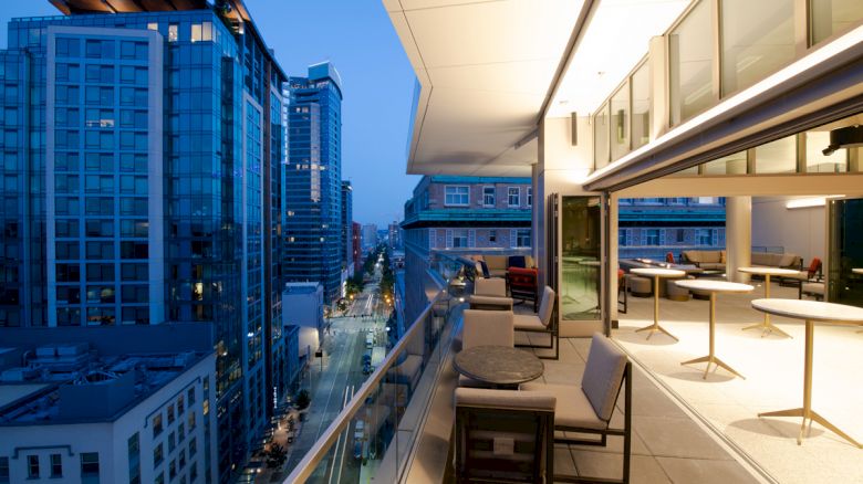 A modern balcony with tables and chairs overlooks a cityscape with tall buildings at dusk under a clear, blue evening sky.