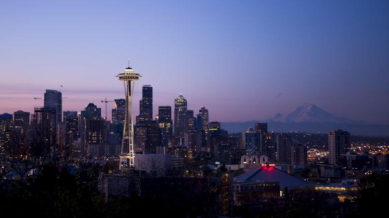 The image shows the Seattle skyline at dusk, featuring the Space Needle and Mt. Rainier in the background against a purple sunset sky.