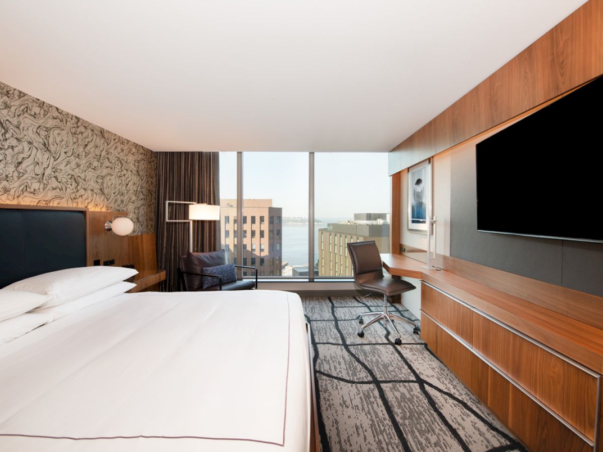 A modern hotel room with a spacious bed, desk, large TV, and floor-to-ceiling windows showing a cityscape view.