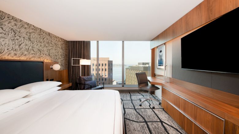 A modern hotel room with a spacious bed, desk, large TV, and floor-to-ceiling windows showing a cityscape view.