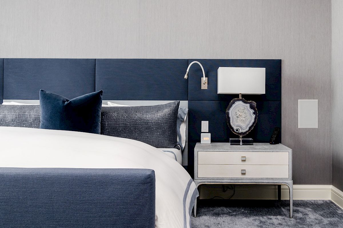 A modern bedroom features a blue upholstered headboard, a white bedside table with a lamp, blue and white bedding, and a gray carpet.